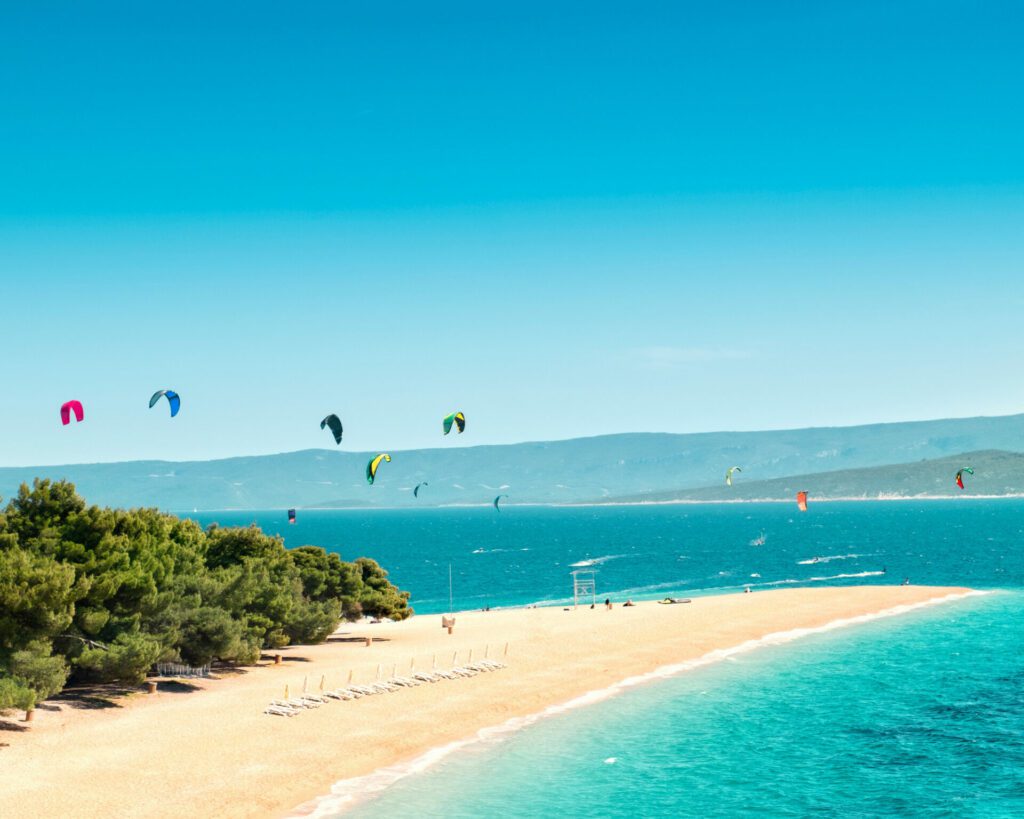 plage blanche avec une forêt au bord de la plage, avec plusieurs voile de kitesurf élevées dans les airs , l'eau est turquoise